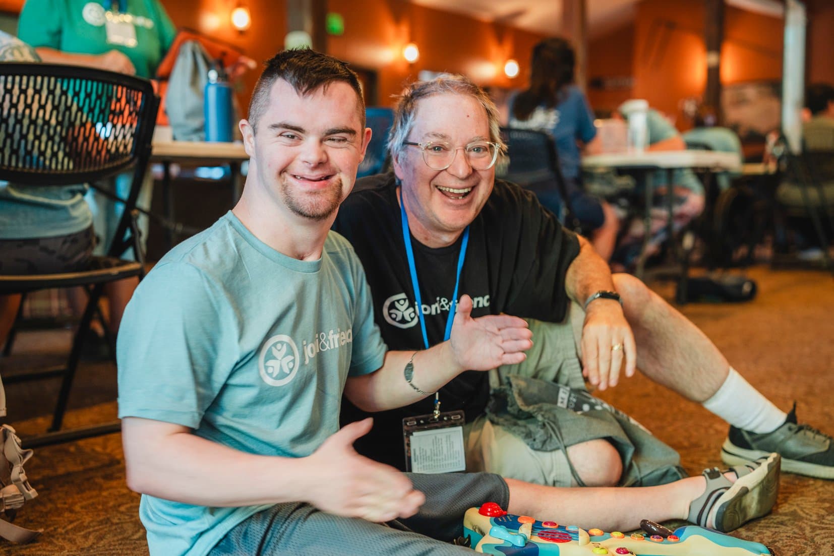 A man with Down syndrome sitting on the floor with another man; both are smiling and posing for the camera.
