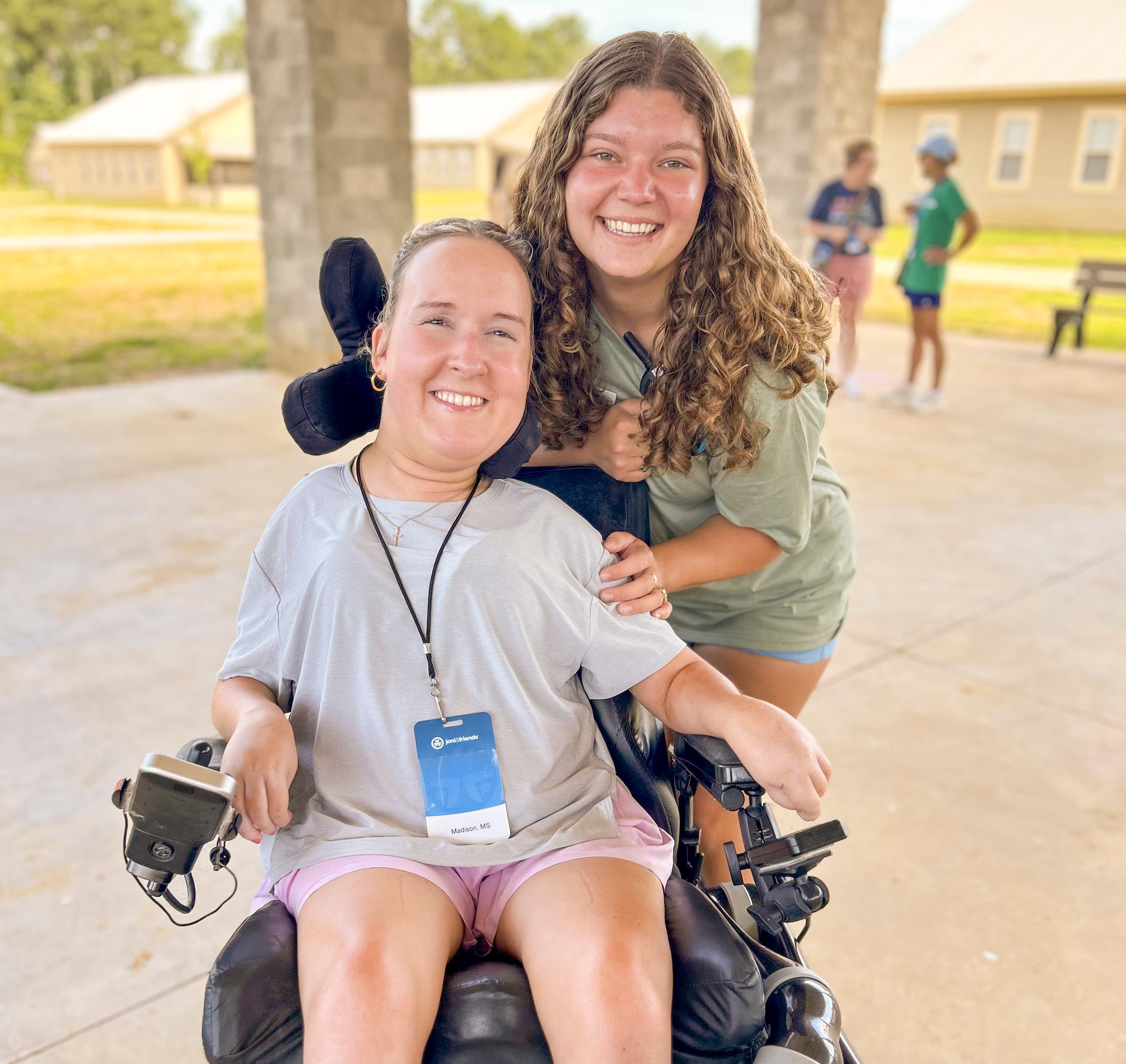 A woman in a wheelchair next to another woman. Both of them are smiling widely for the camera.