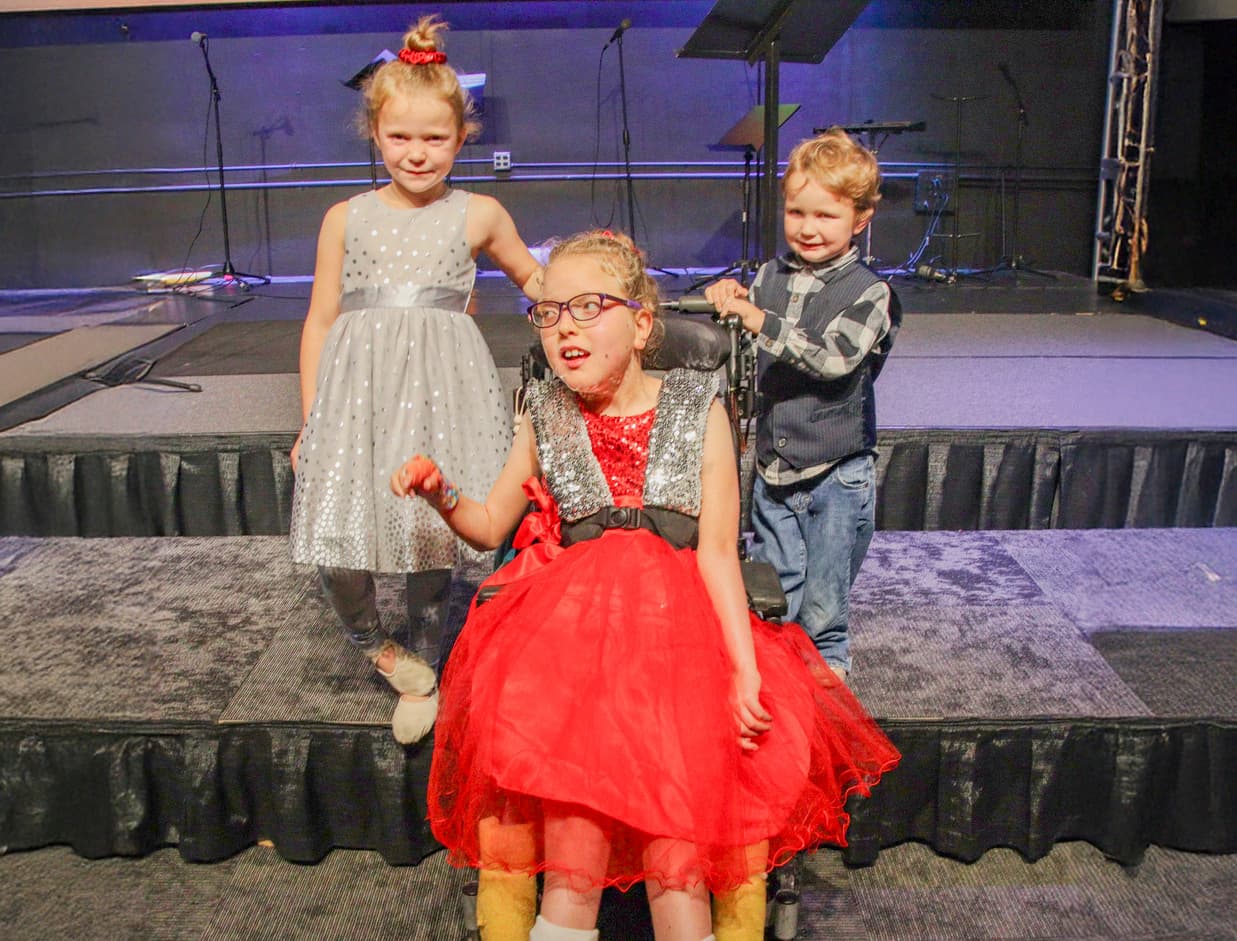 A girl in a beautiful dress sitting in a wheelchair, smiling at the camera with her two younger siblings.