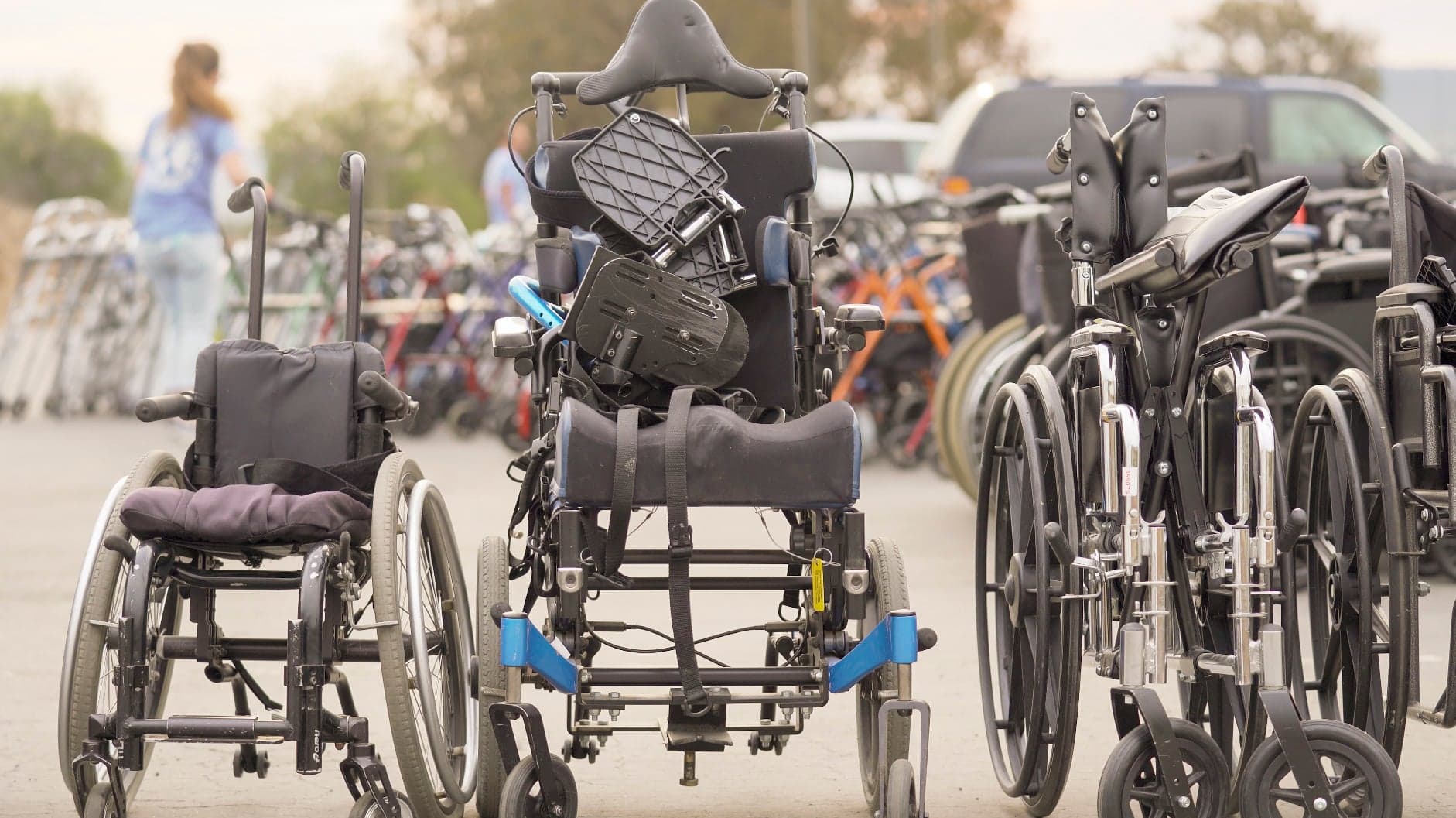 A front-facing manual wheelchair is parked outdoors on a paved lot alongside several folded wheelchairs and walkers. A blurred person stands in the background.