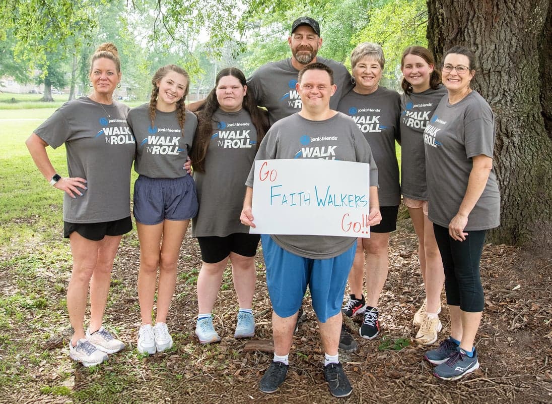 A man with Down syndrome holds a cardboard sign reading “Go Faith Walker Go!!” while people stand around him.