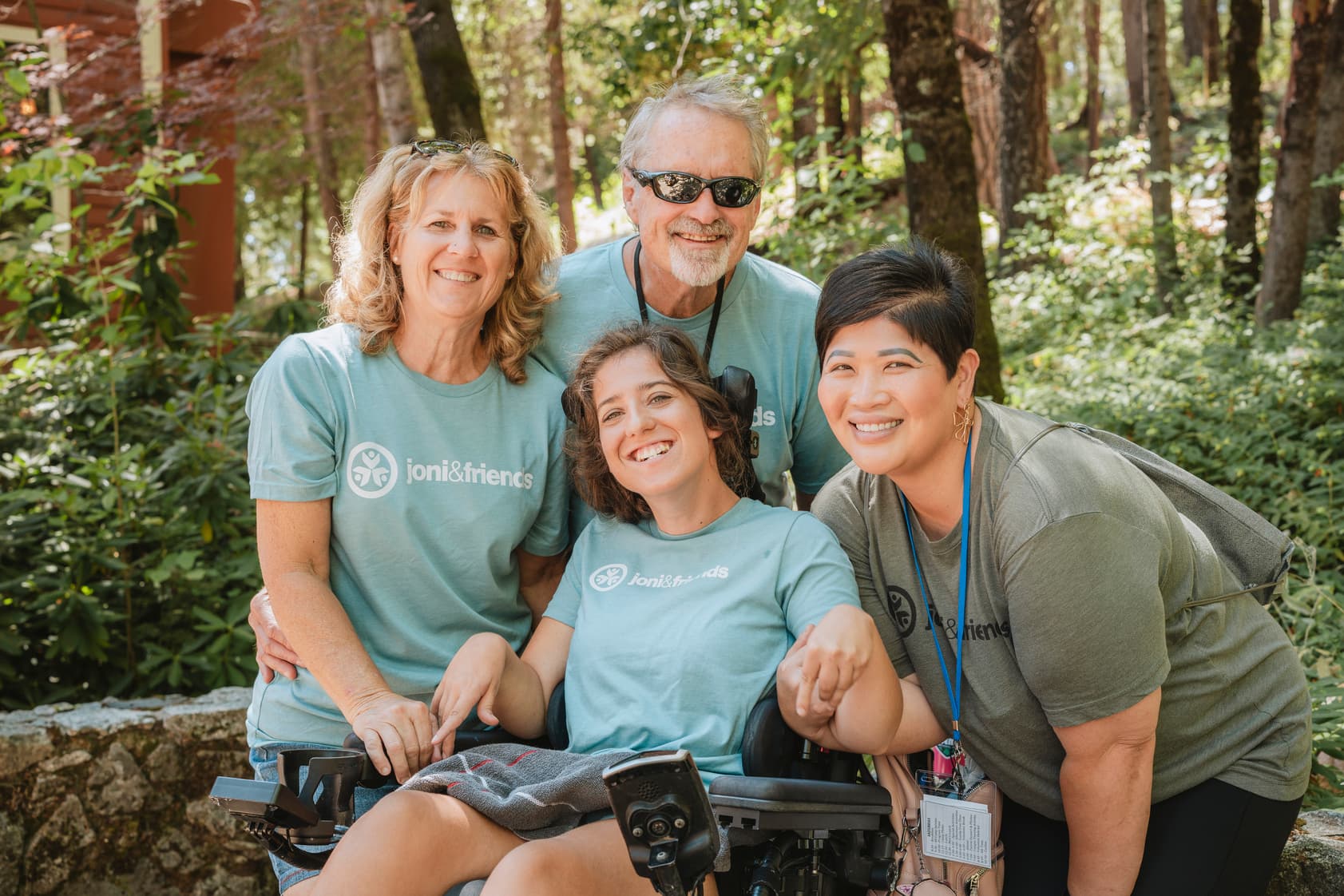 Woman in a wheelchair with her parents and a volunteer, outdoors among trees and plants.