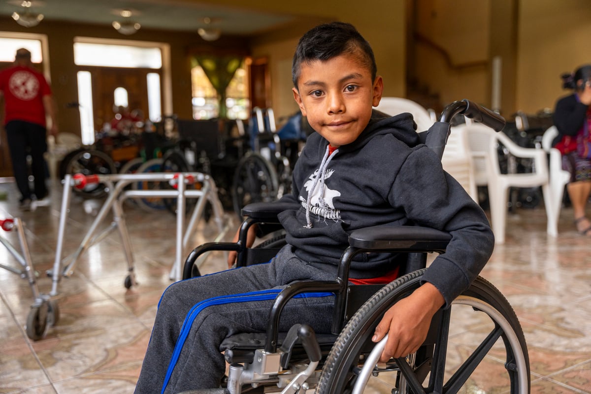 Jason wearing a black sweater and dark grey pants sitting in his new wheelchair posing and smiling for the camera. 