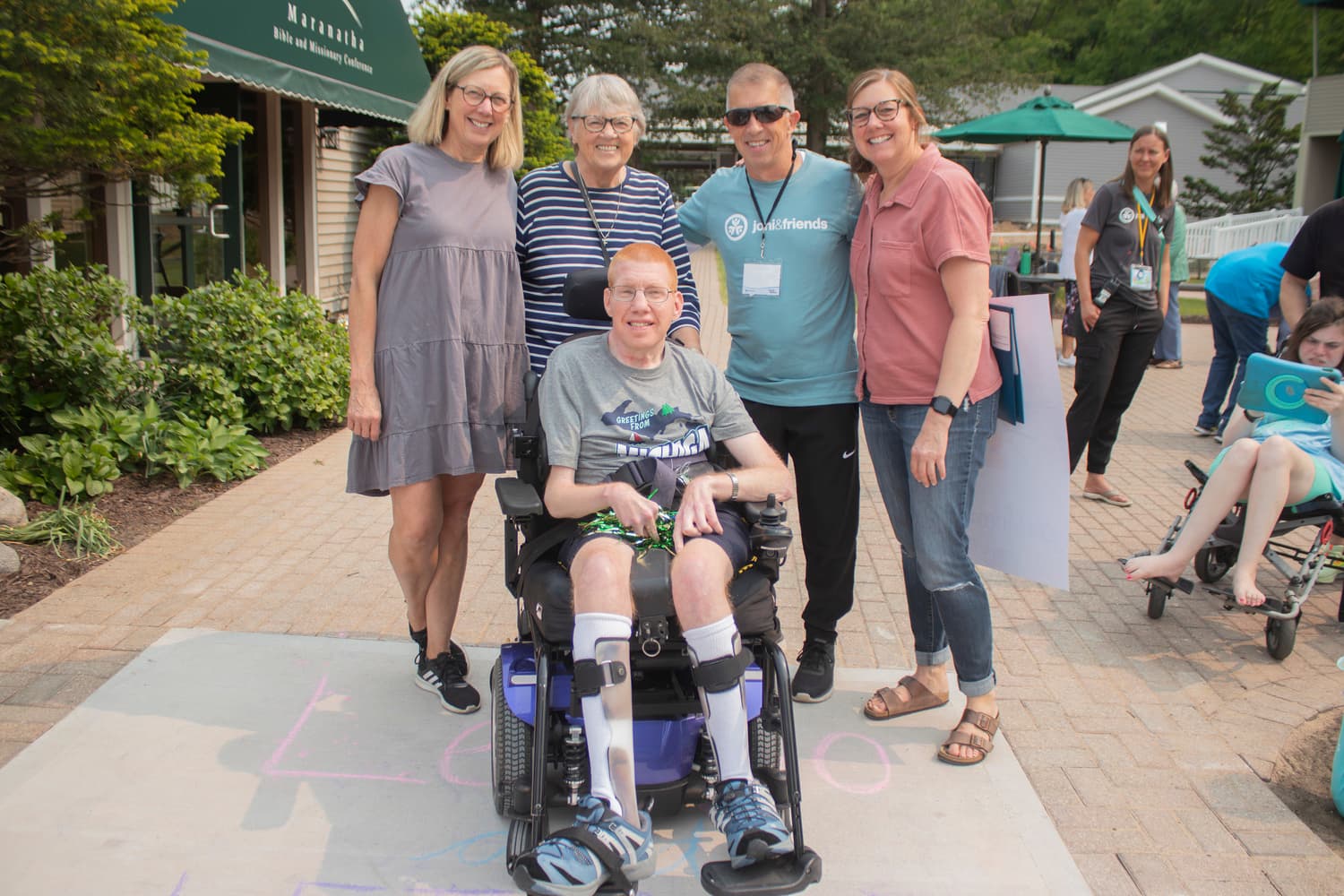 A man in a power wheelchair poses with four people standing behind him; others are visible in the background, all smiling at the camera.