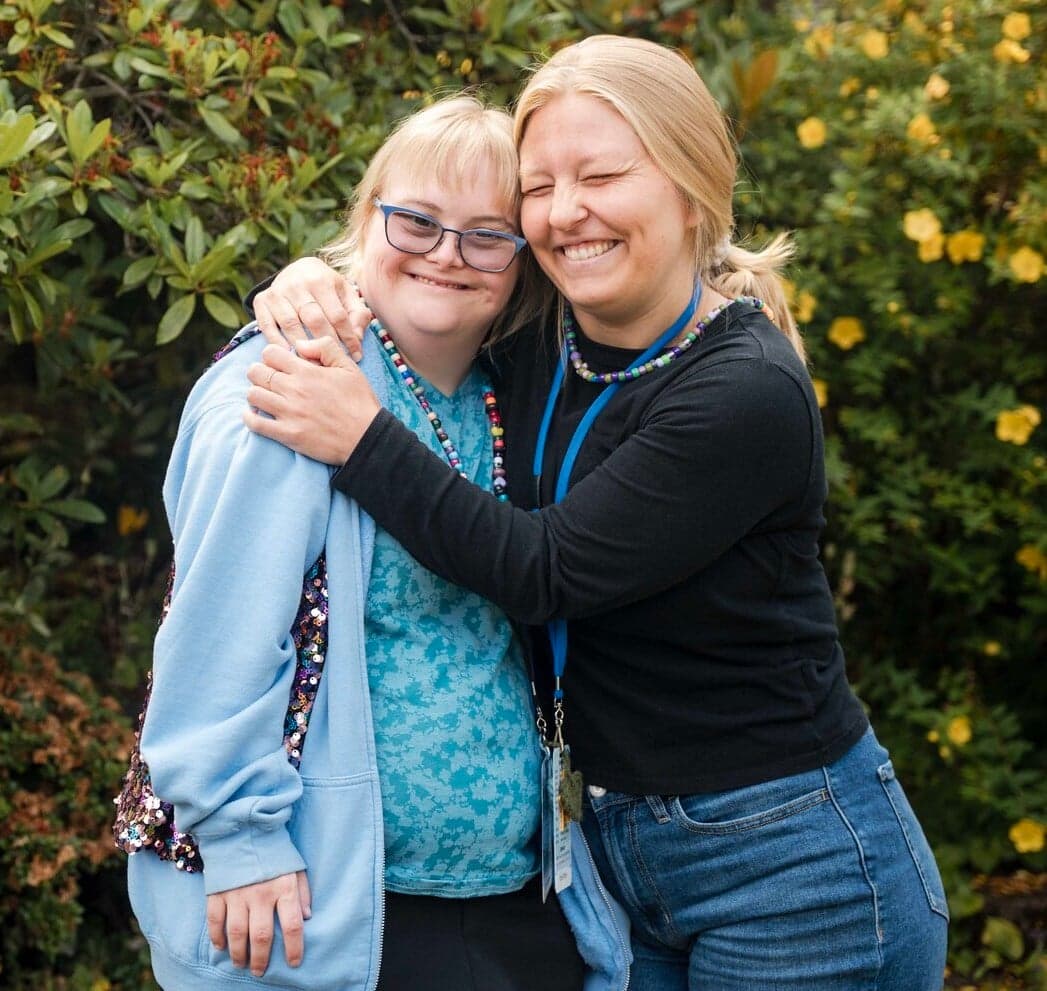 A woman hugging a woman with Down Syndrome. Both are smiling for the camera. 