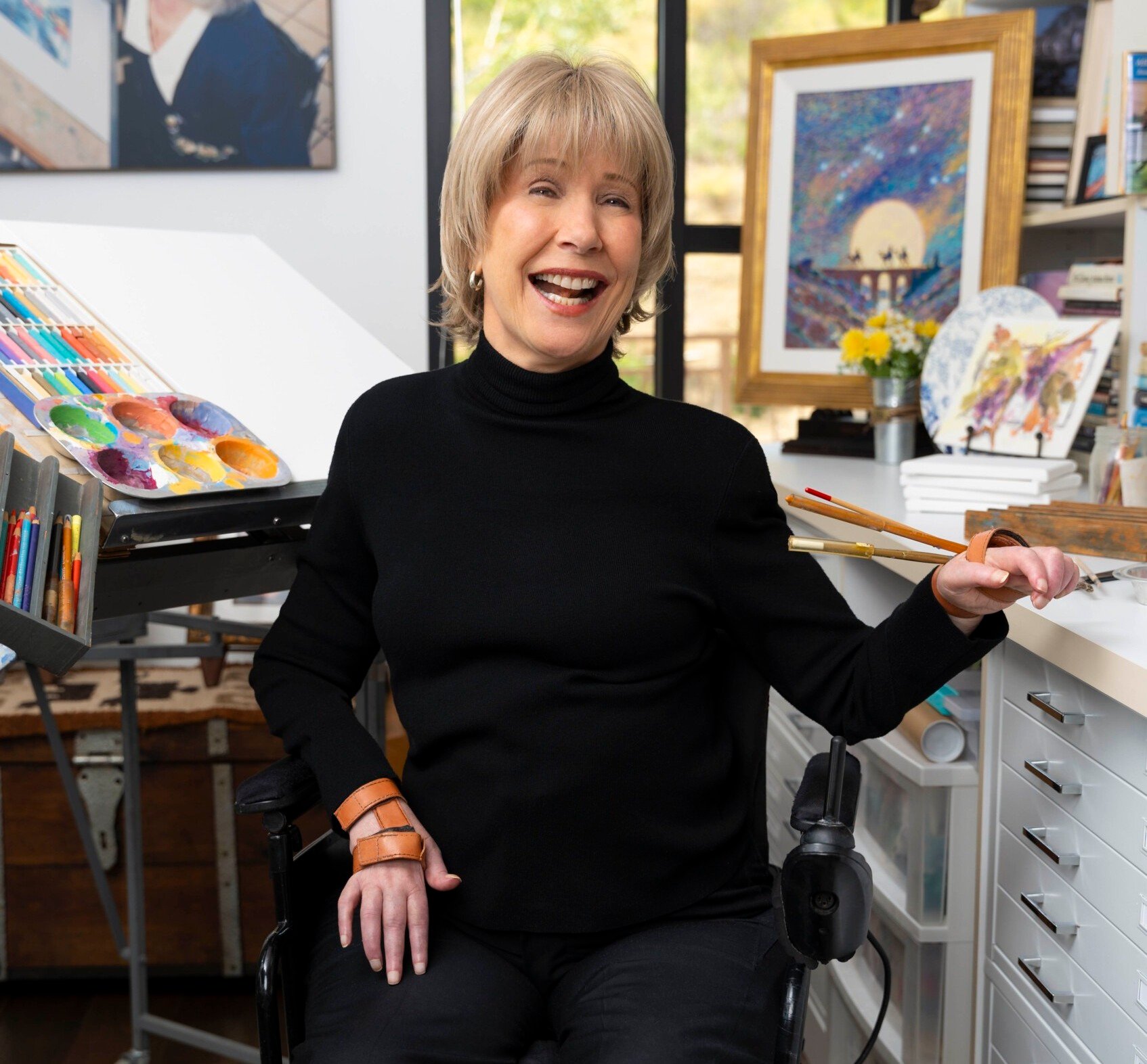 Joni smiling and posing in her art room, with her artwork and art supplies displayed in the background.