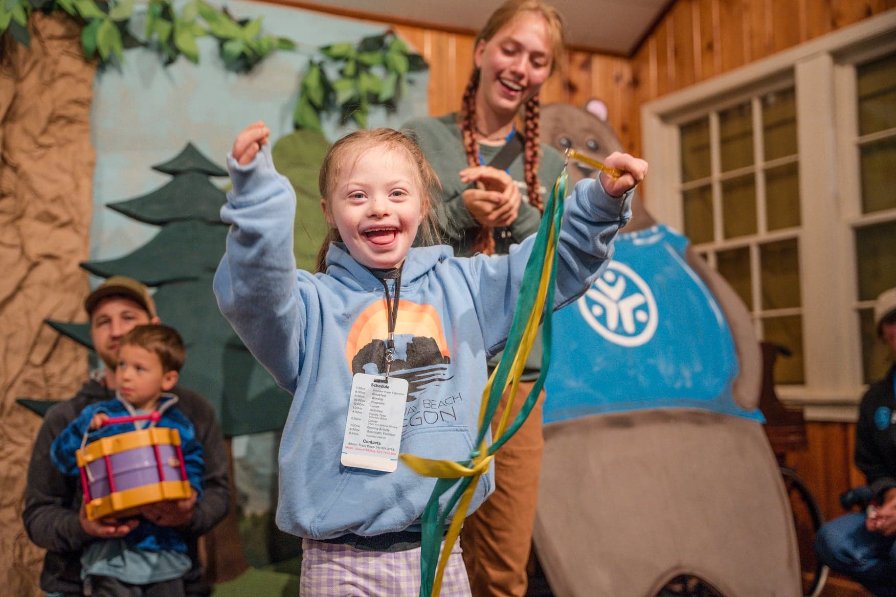 A young girl waves a ribbon while smiling, with people in the background.