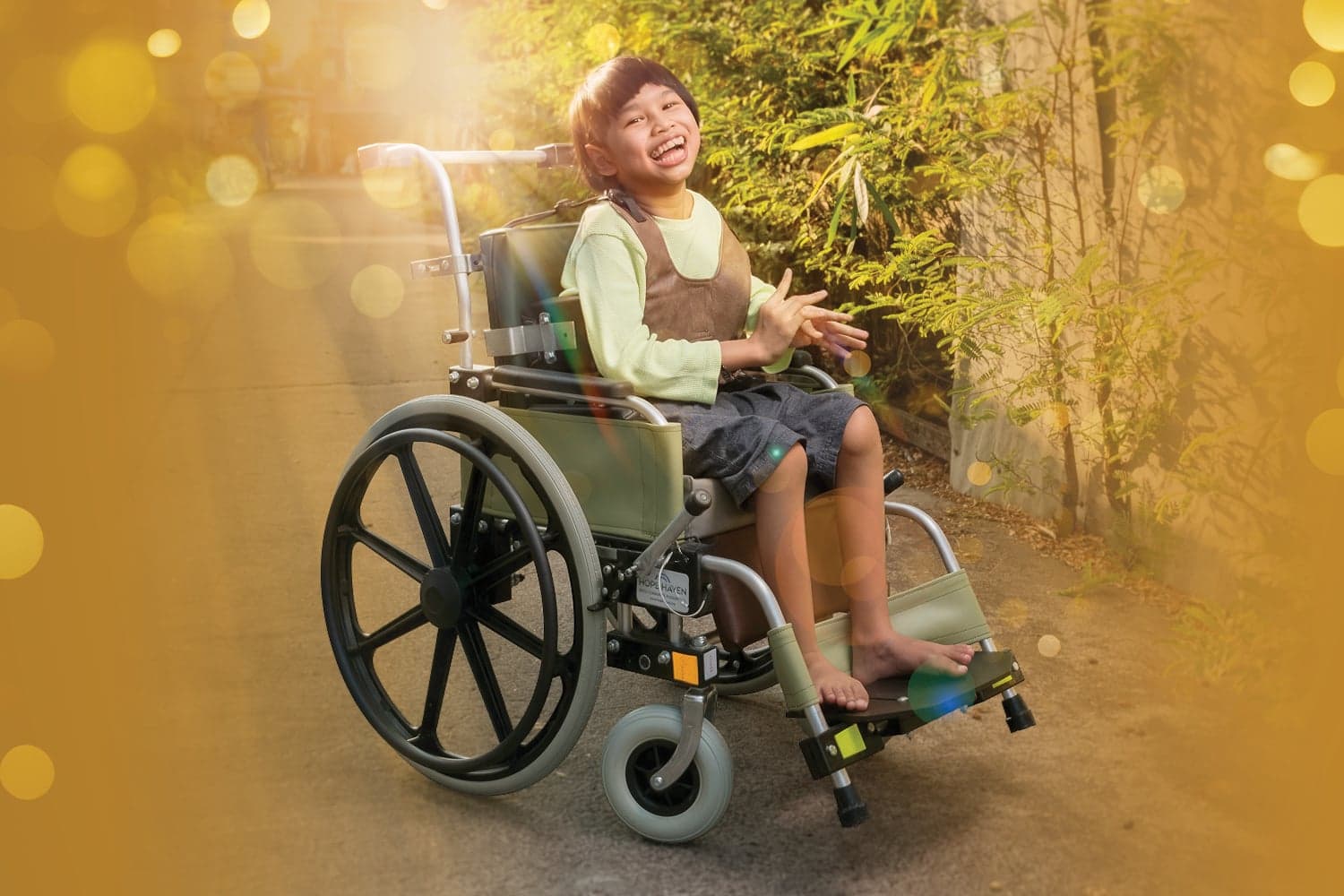 A little girl smiles joyfully in her new wheelchair outdoors, with a softly blurred, bokeh-effect background.