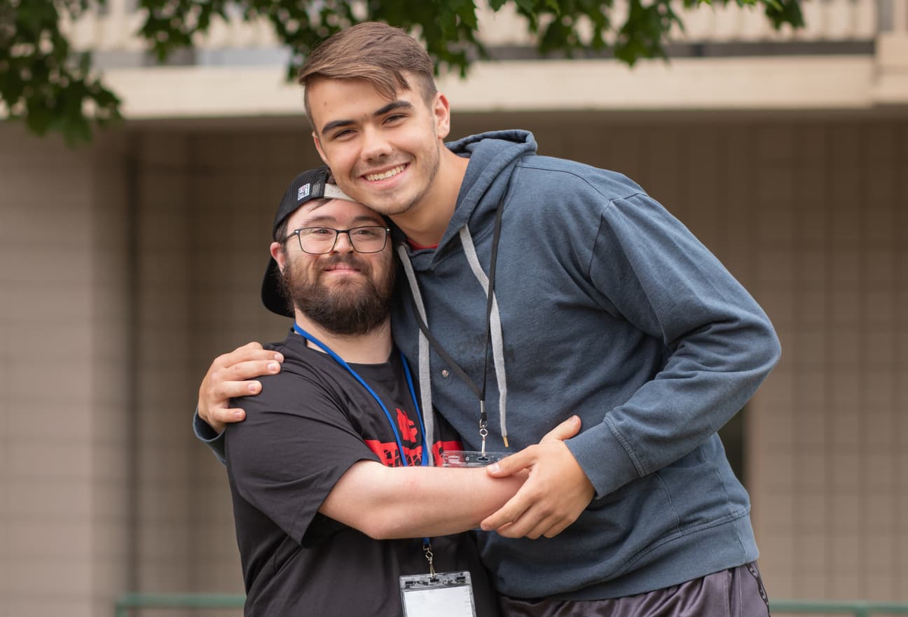 A man with Down syndrome stands next to another man. Both are smiling and posing for the camera.