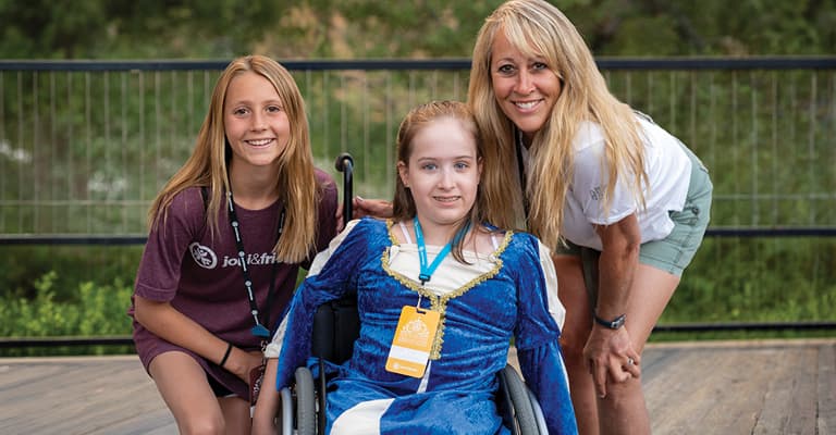 Two smiling volunteers kneel beside a girl in a wheelchair at an outdoor event on a wooden deck, all wearing lanyards.